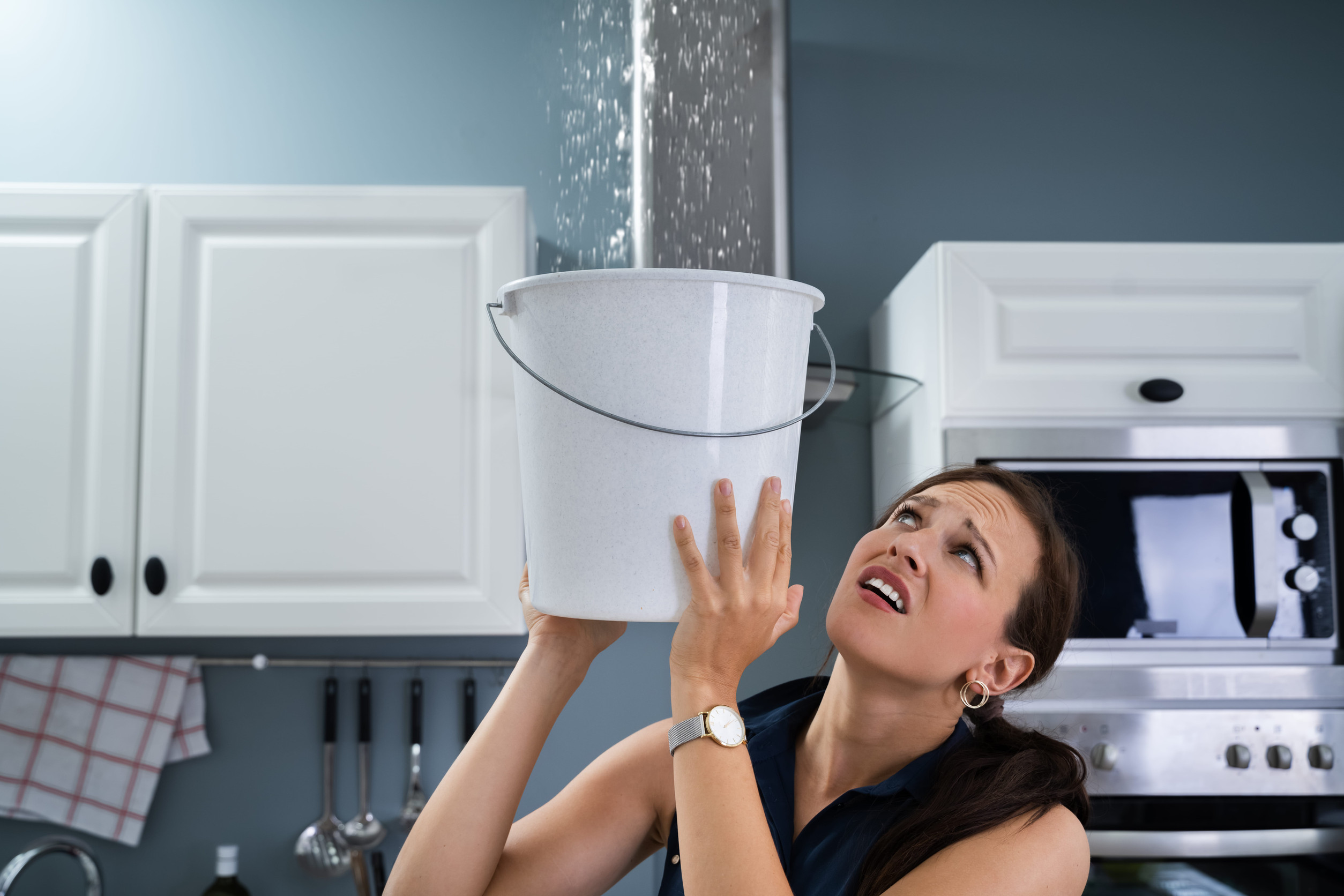 A woman holds a bucket under a leaking ceiling in a kitchen, looking up worriedly as water pours down—likely needing storm damage repair or even roof replacement—with white cabinets and a microwave visible in the background.