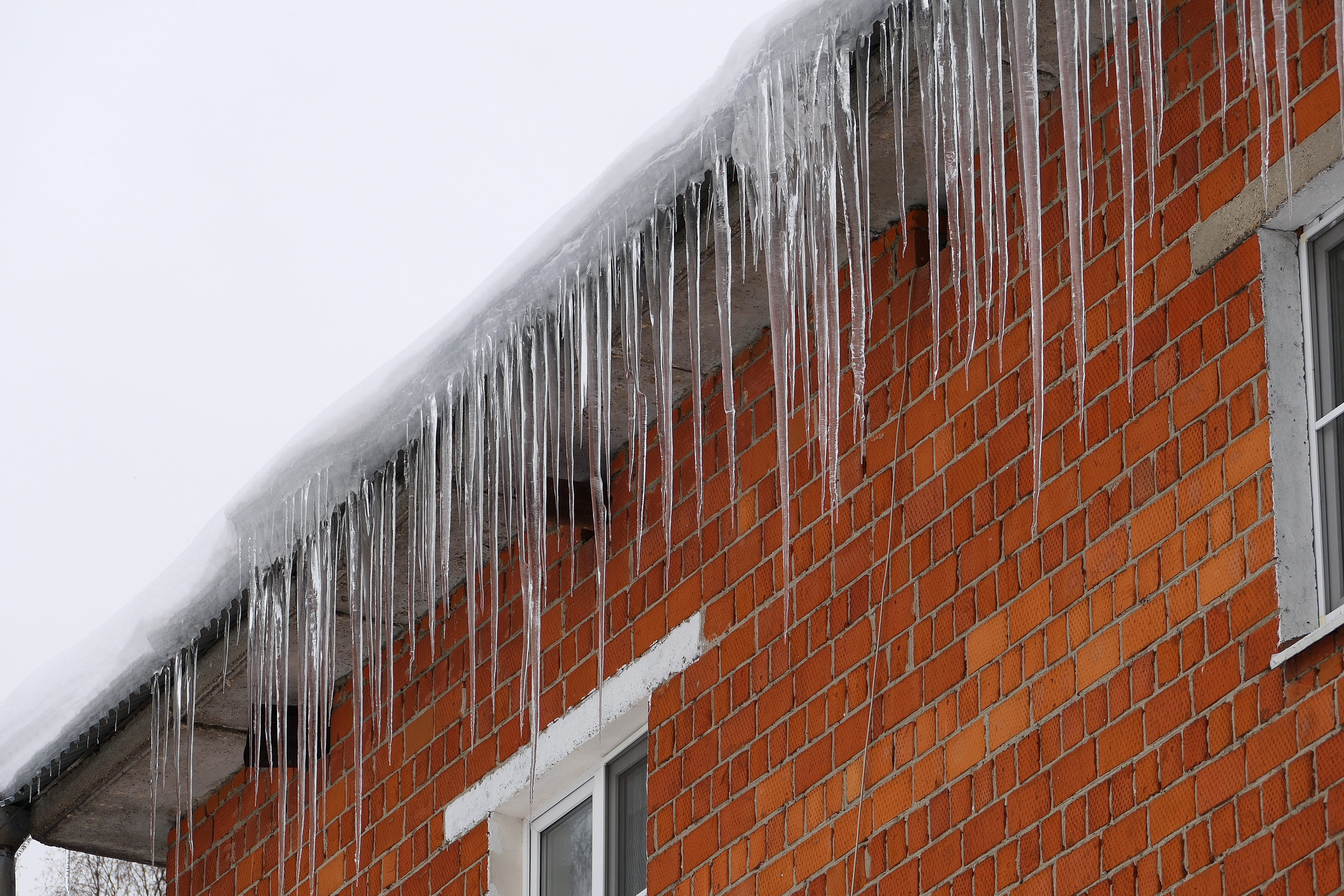 Long icicles hang from the snowy roof of a red brick building with white window frames on a cloudy winter day.