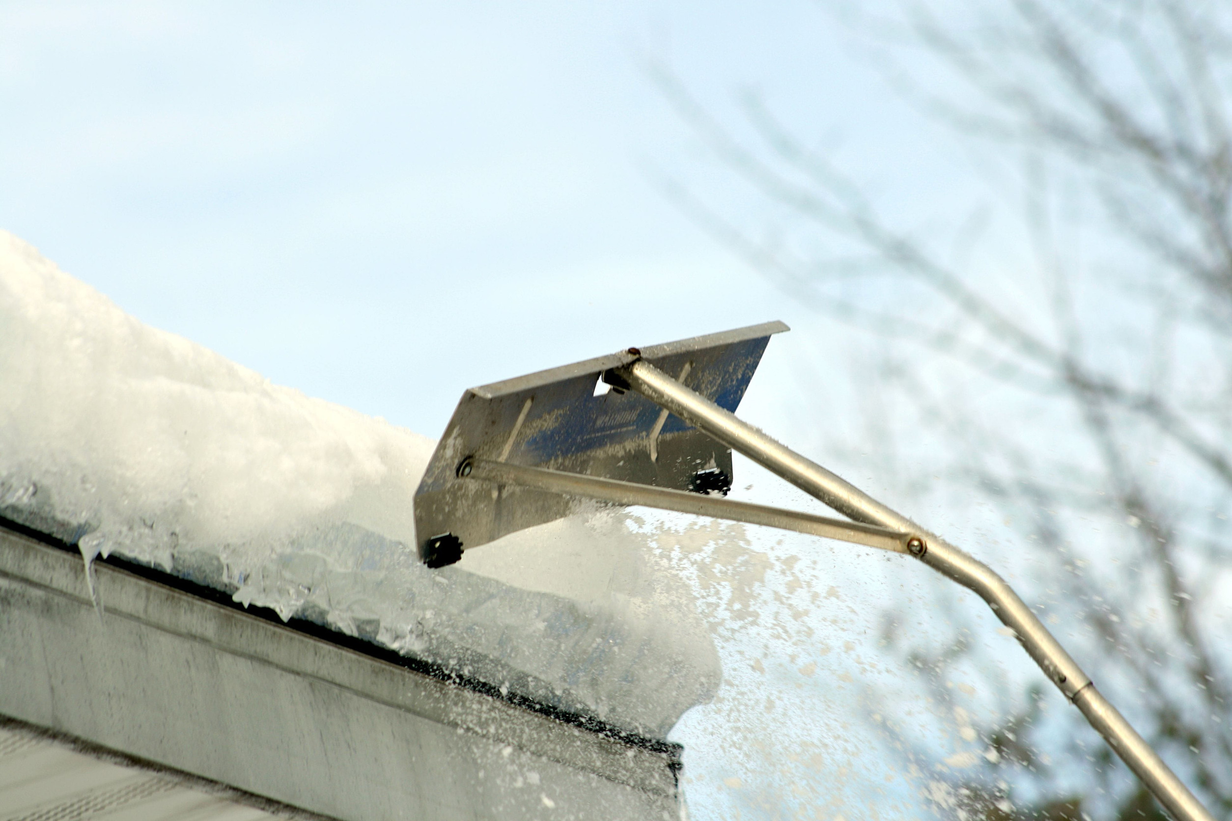A person uses a roof rake to clear snow and ice from a rooftop.
